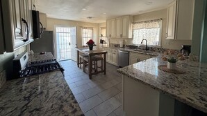 Une cuisine dotée de plans de travail en marbre, un coin repas avec une table et des chaises, et une vue sur l’extérieur par une porte.