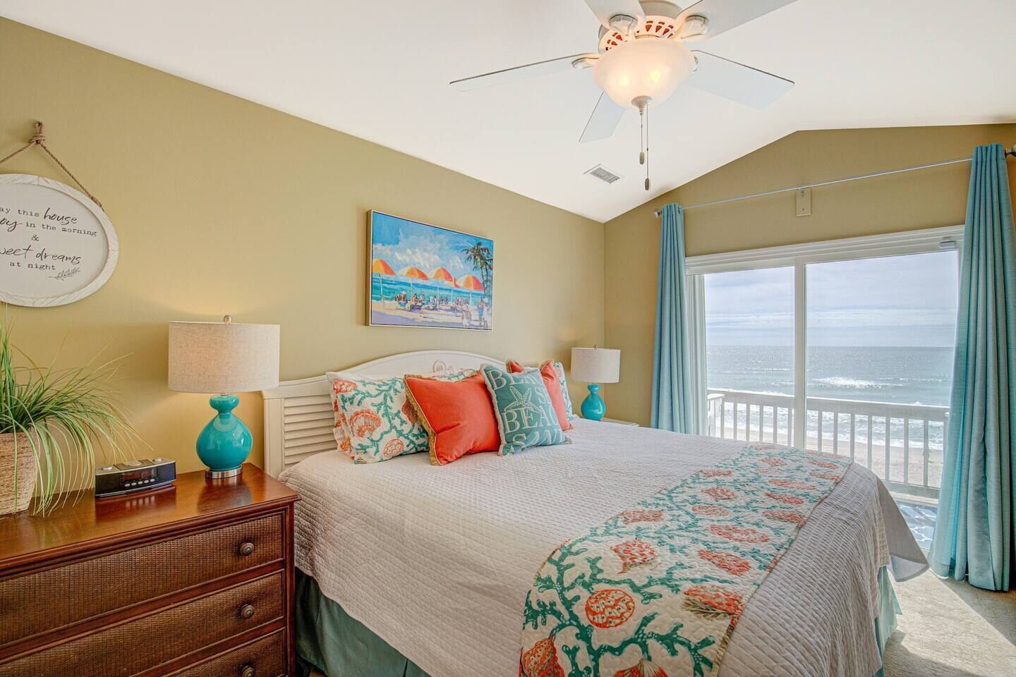A bedroom with a bed, two bedside tables, a nightstand, a ceiling fan, a window with curtains, and a view of the ocean.