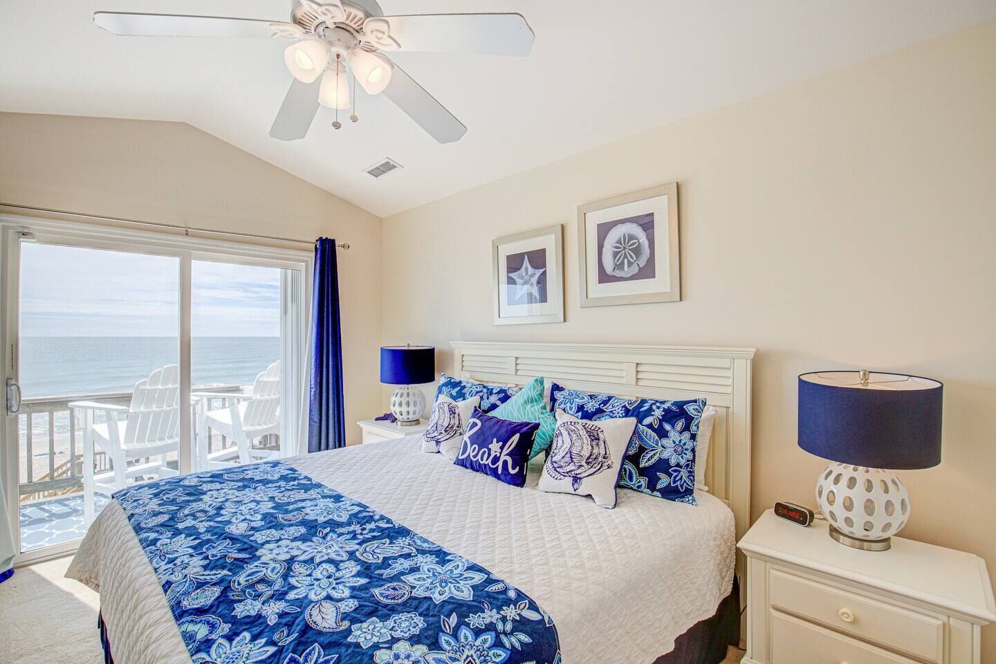 A bedroom with a bed, a nightstand, a lamp, a ceiling fan, and a view of the ocean through a sliding door.