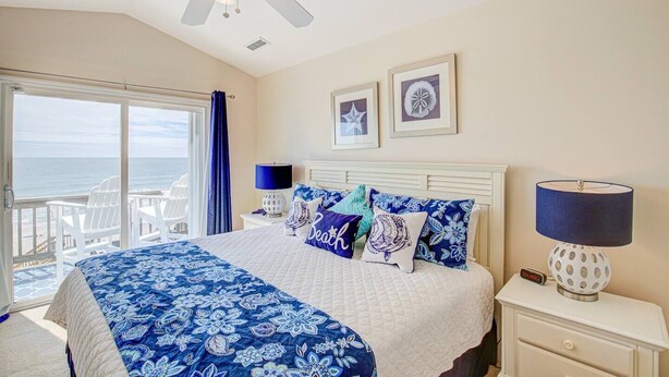 A bedroom with a bed, a nightstand, a lamp, a ceiling fan, and a view of the ocean through a sliding door.