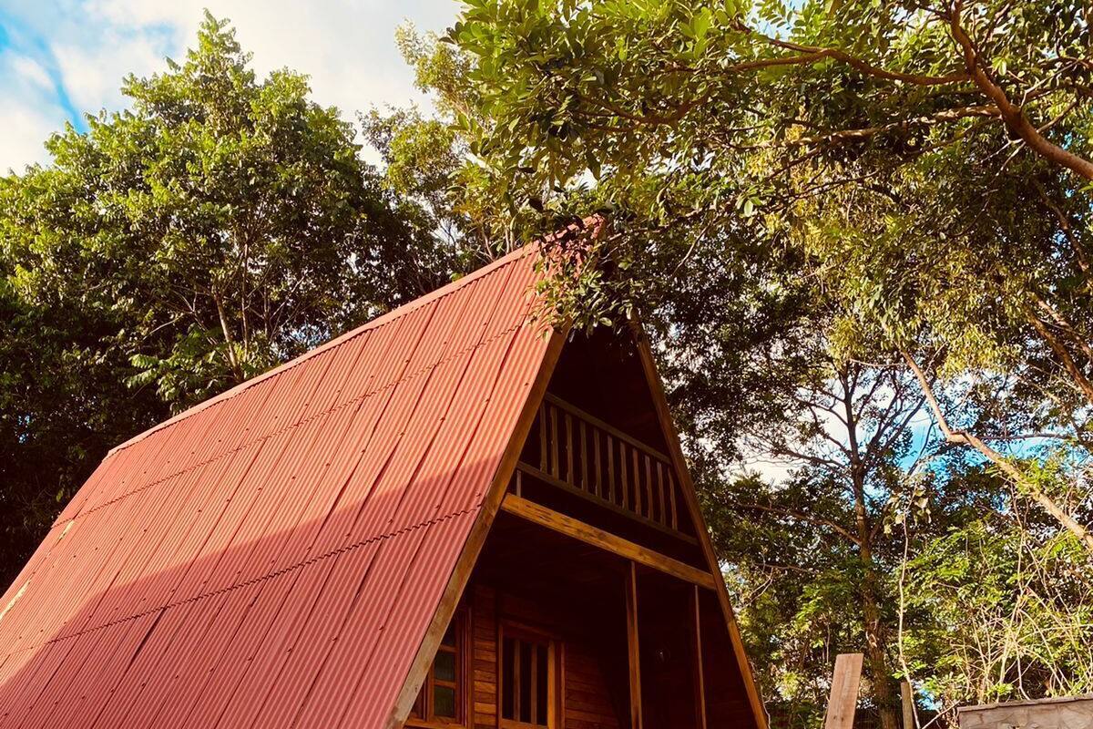 A rustic wooden cabin with a sloped red roof, stone walls, and a wooden porch with steps leading to a door.