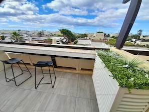 A rooftop terrace with two bar stools, a planter with greenery, and a view of the cityscape.