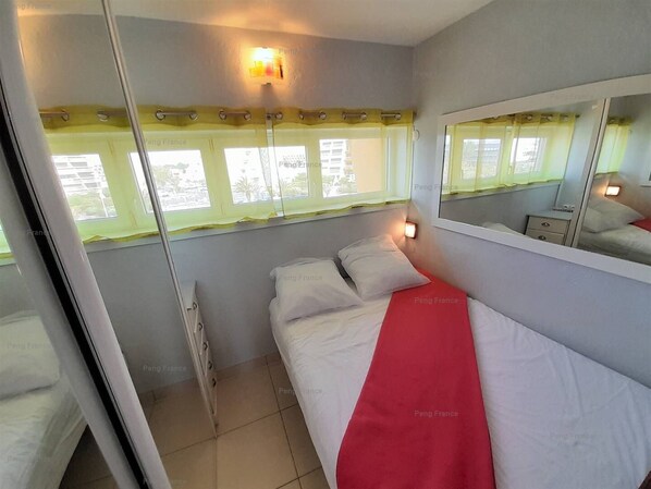 Un dormitorio con cama, mesita de noche, espejo y ventana con cortinas.