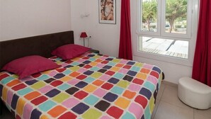 Een slaapkamer met een kleurrijk beddegoed, een raam met rode gordijnen en een witte muur met een ingelijst beeld.