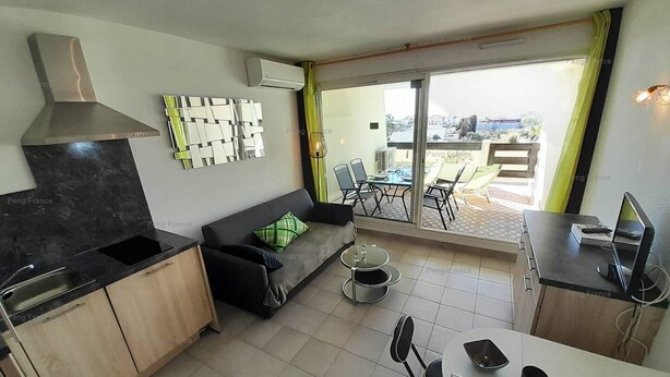 Cozinha moderna com bancada preta, armários de madeira e fogão embutido. Há uma área de jantar com mesa e cadeiras, e uma varanda com vista.
