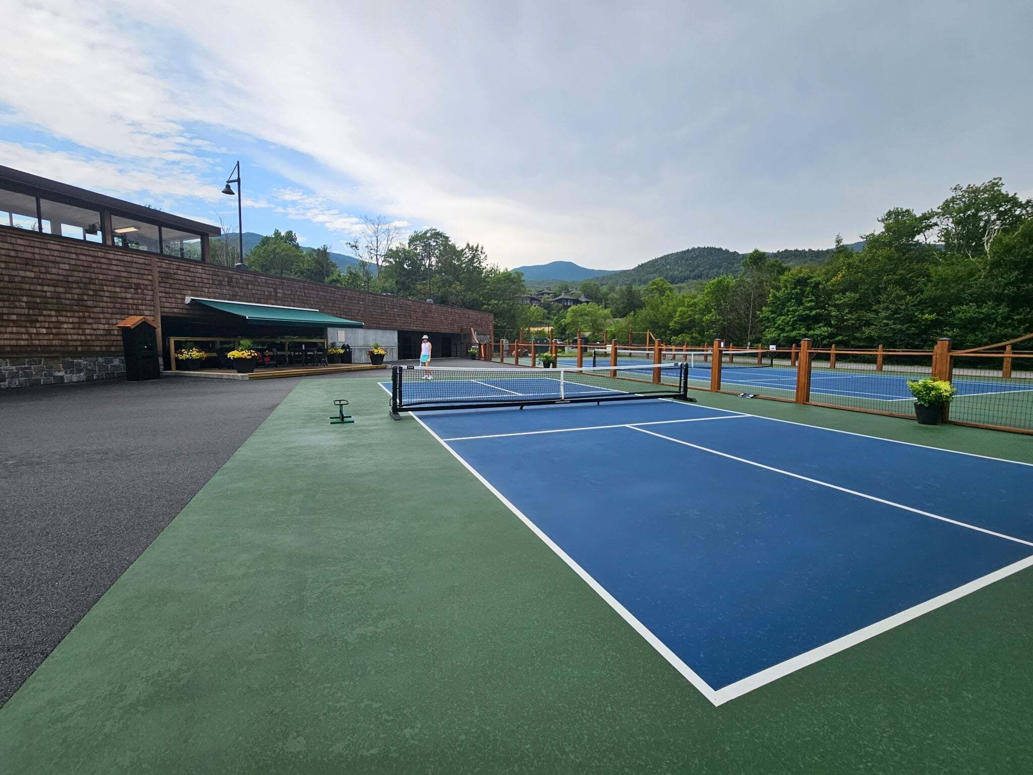 An outdoor tennis court with a net, surrounded by a wooden fence, and a building with a green awning in the background.