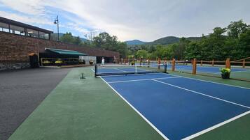 An outdoor tennis court with a net, surrounded by a wooden fence, and a building with a green awning in the background.