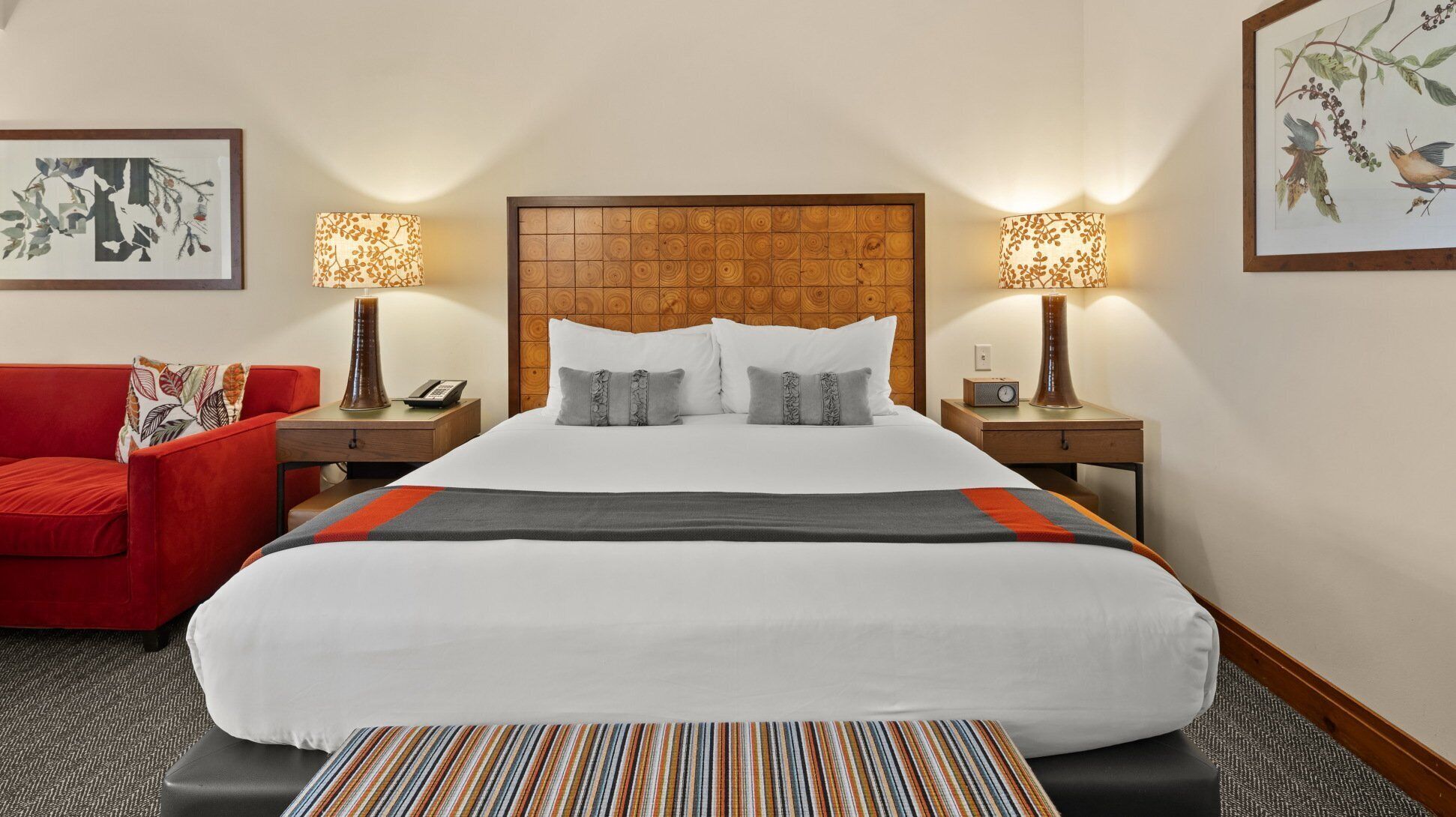Habitación de hotel con una cama grande, dos mesitas de noche con lámparas, un sillón rojo y dos cuadros en las paredes.