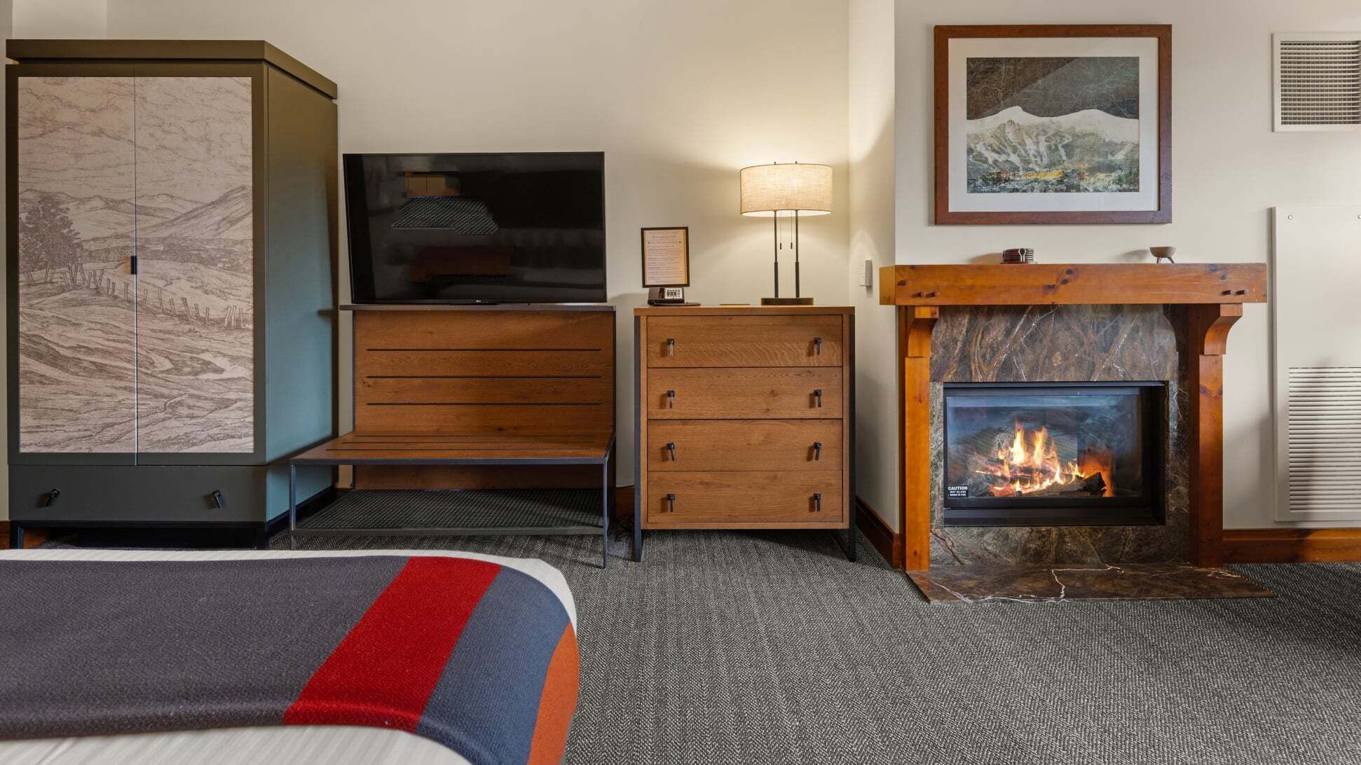 A hotel room with a bed, a television, a desk with drawers, a lamp, a fireplace, and a cabinet with a map.