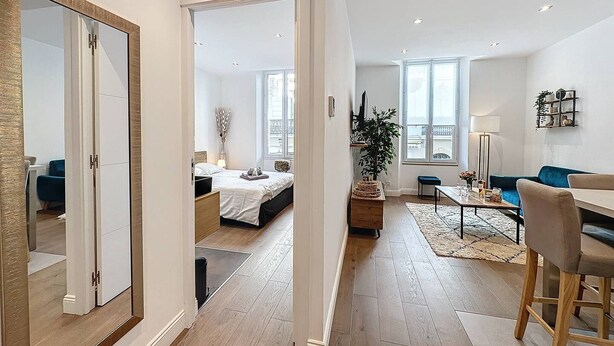 Un apartamento moderno con un espejo grande, un comedor con mesa y sillas, una cama con mesita de noche y un ventanal que ofrece luz natural.