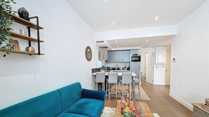 Un apartamento moderno con un sofá color turquesa, una mesa de centro de madera con flores, una cocina con armarios grises y una puerta blanca que da a otra habitación.
