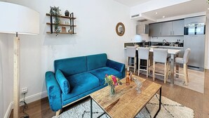 Una sala de estar moderna con un sofá azul, una mesa de centro con flores, y una cocina al fondo.