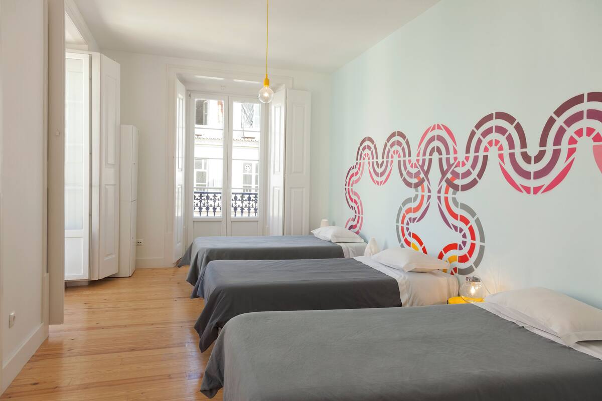 A hotel room with two beds, a wooden floor, a light-colored wall with a decorative design, and a window with a view of the outside.
