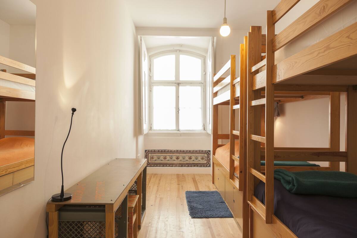 A dormitory room with bunk beds, a desk, a lamp, and a window with a view.