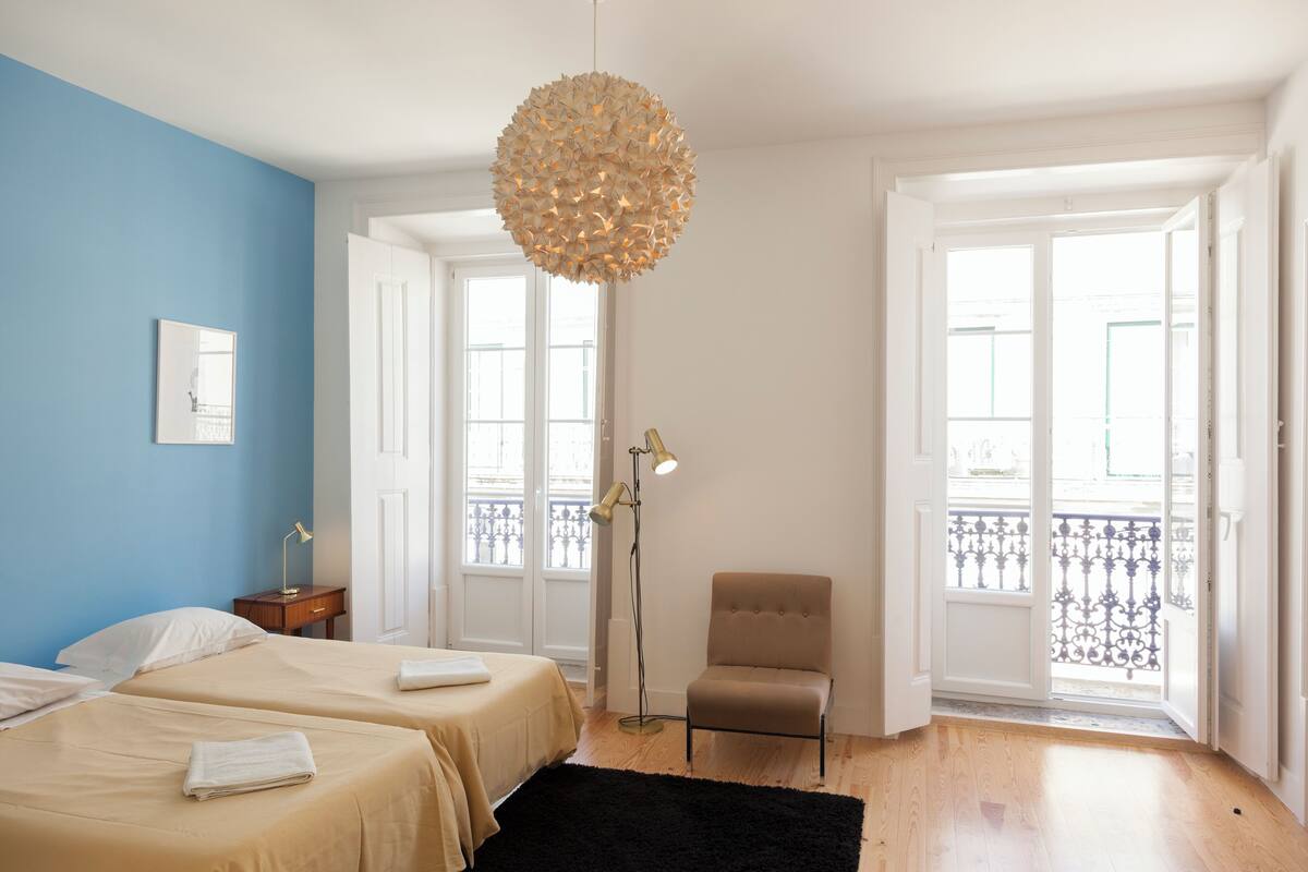 A hotel room with two beds, a chair, a floor lamp, and a large window with a balcony view.