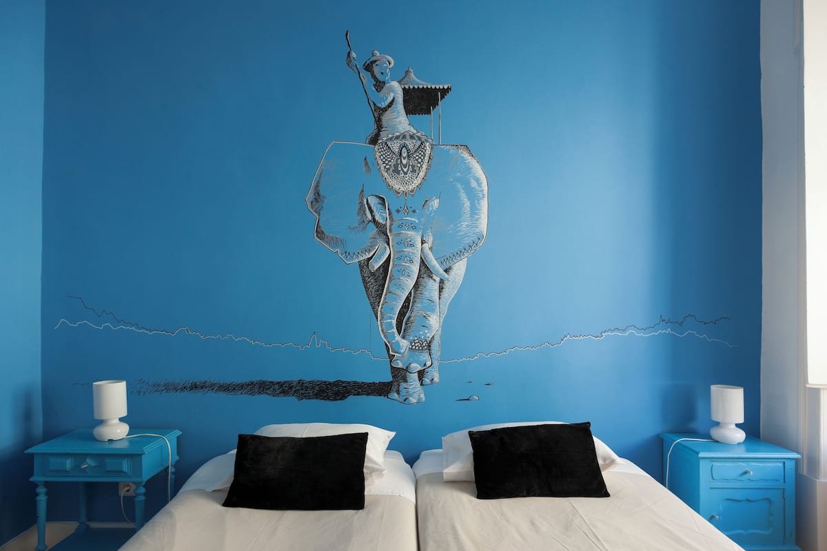 A bedroom with two beds, a mural of a person on an elephant, and a blue wall.