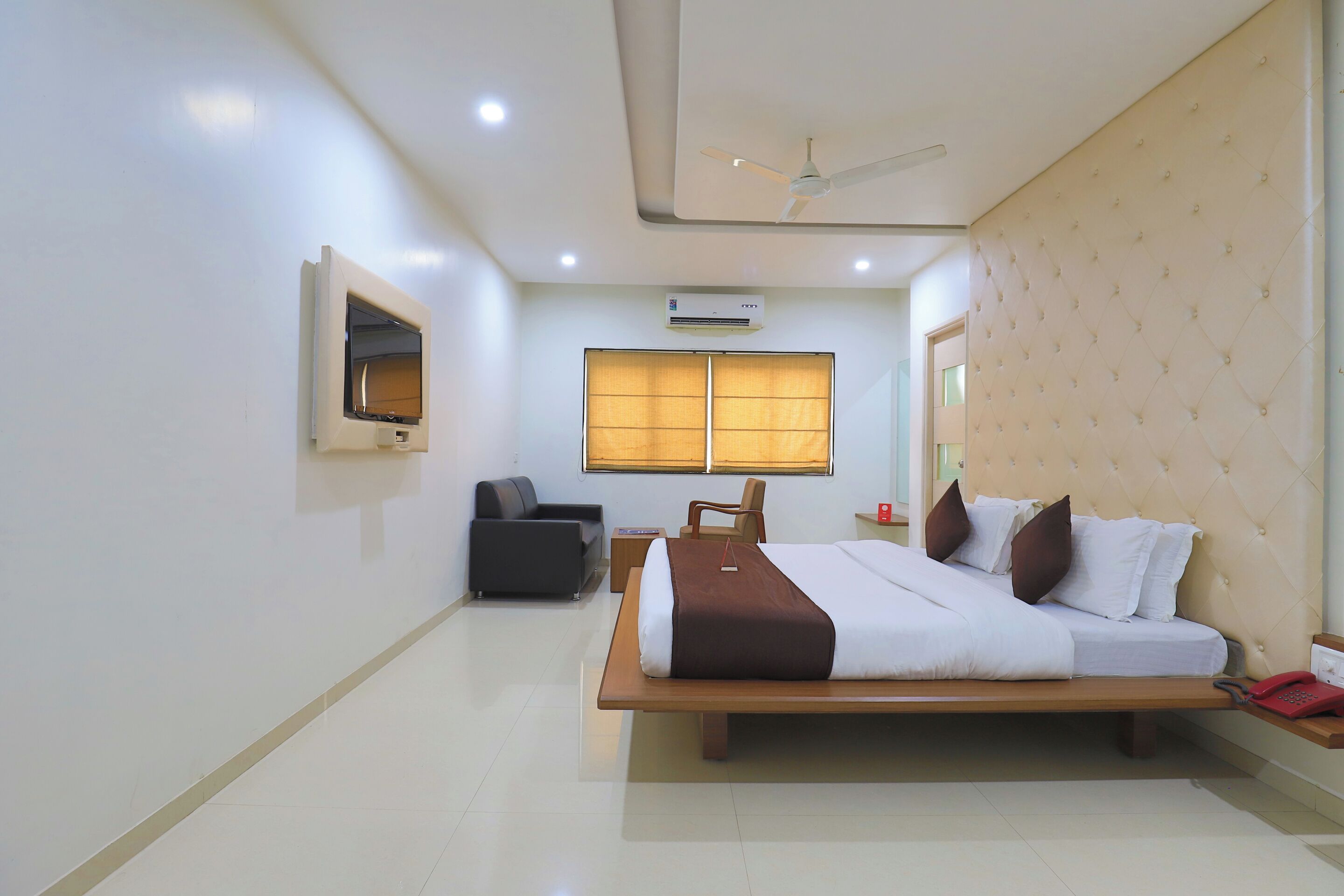 A hotel room with a bed, a chair, a small table, a mirror, and a ceiling fan.