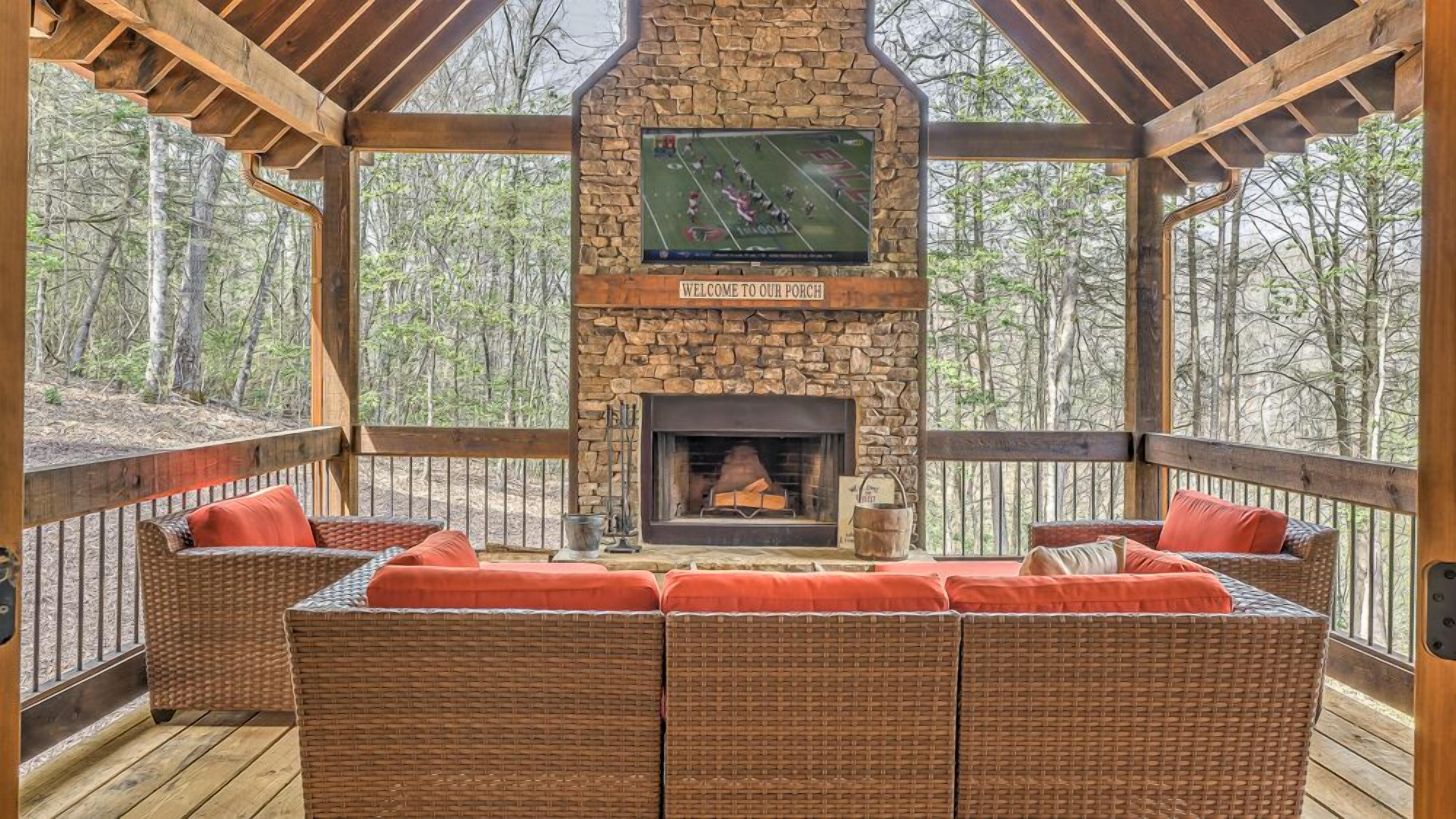 A covered outdoor seating area with a stone fireplace, a television mounted above it, and a view of the surrounding forest.