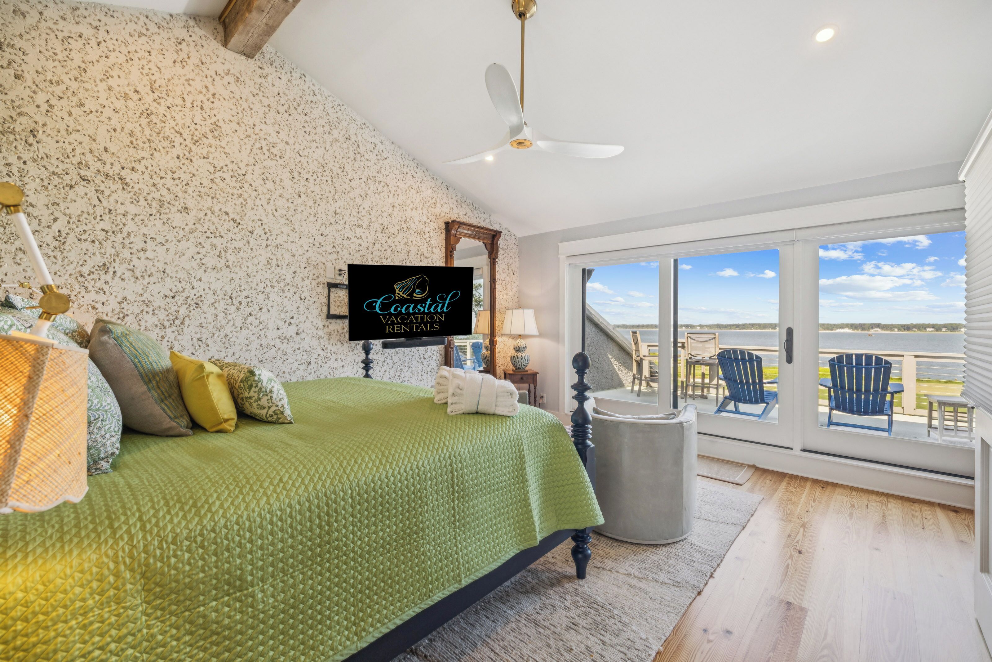 A bedroom with a green quilted bedspread, a ceiling fan, and a large window offering a view of the beach.