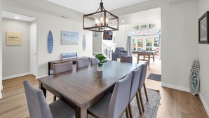 Uma sala de jantar com uma mesa de madeira e cadeiras cinzas, uma luminária pendente e um prancha de surf na parede.