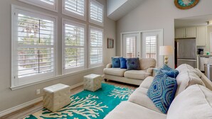 A spacious living room with a beige sofa, patterned cushions, a teal rug, and large windows with blinds. - Walk to the Beach, Shared Heated Pool, Golf Cart Access (Port Aransas)