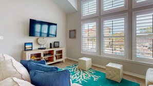 A living room with a TV, wooden cabinet, and ottomans. - Walk to the Beach, Shared Heated Pool, Golf Cart Access (Port Aransas)