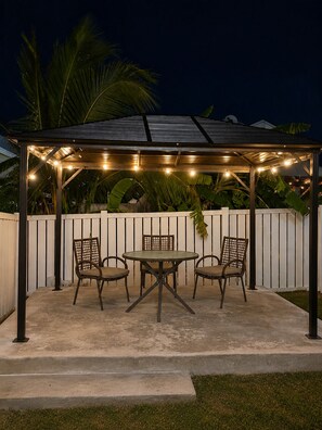 A covered patio area with a table and chairs, string lights, and a white fence in the background.