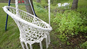 Una hamaca de macramé blanco colgada entre árboles en un área con hierba, con sillas de plástico blancas al fondo.