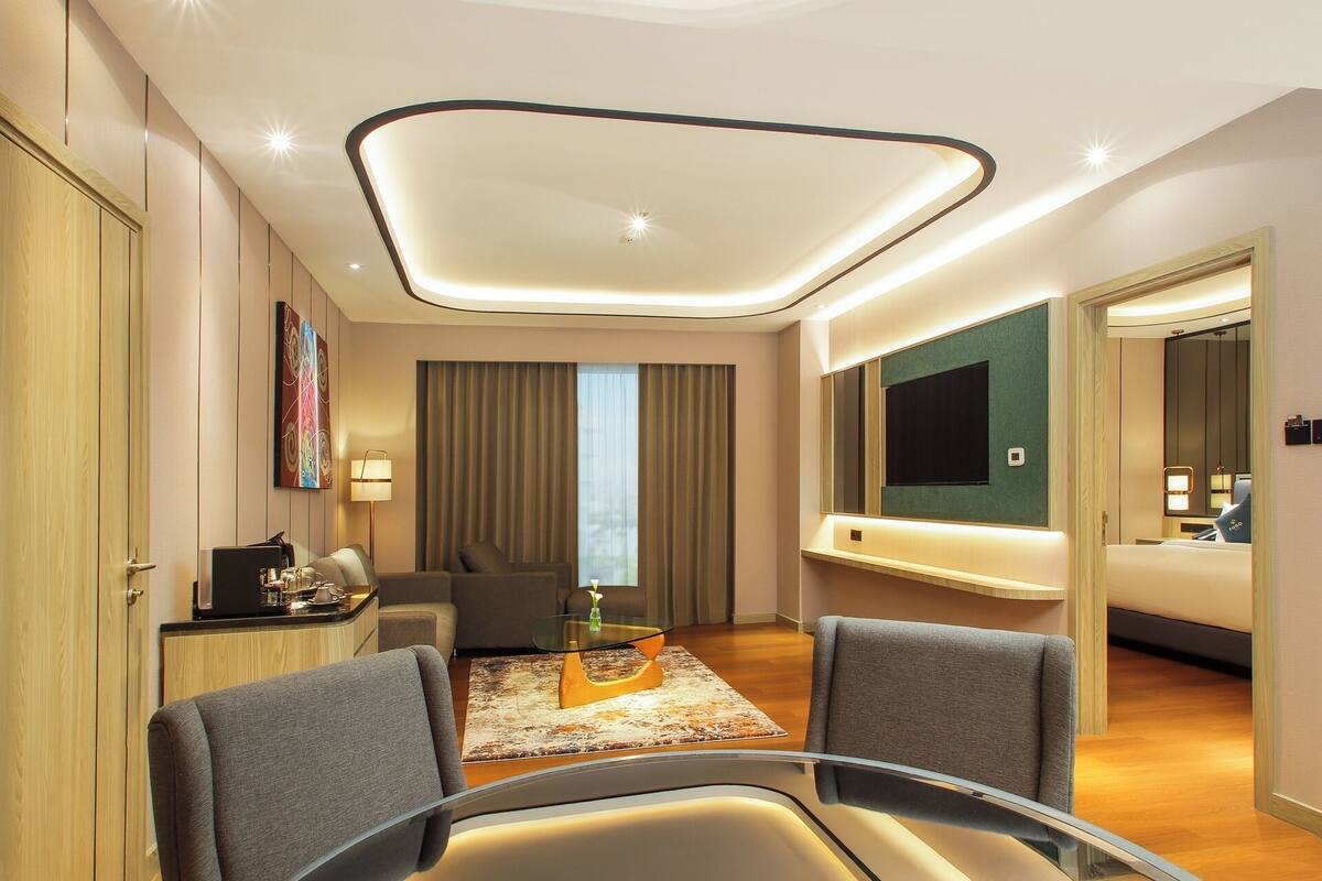A modern hotel room with a glass dining table, two armchairs, a sofa, a TV mounted on the wall, and a large window with curtains.