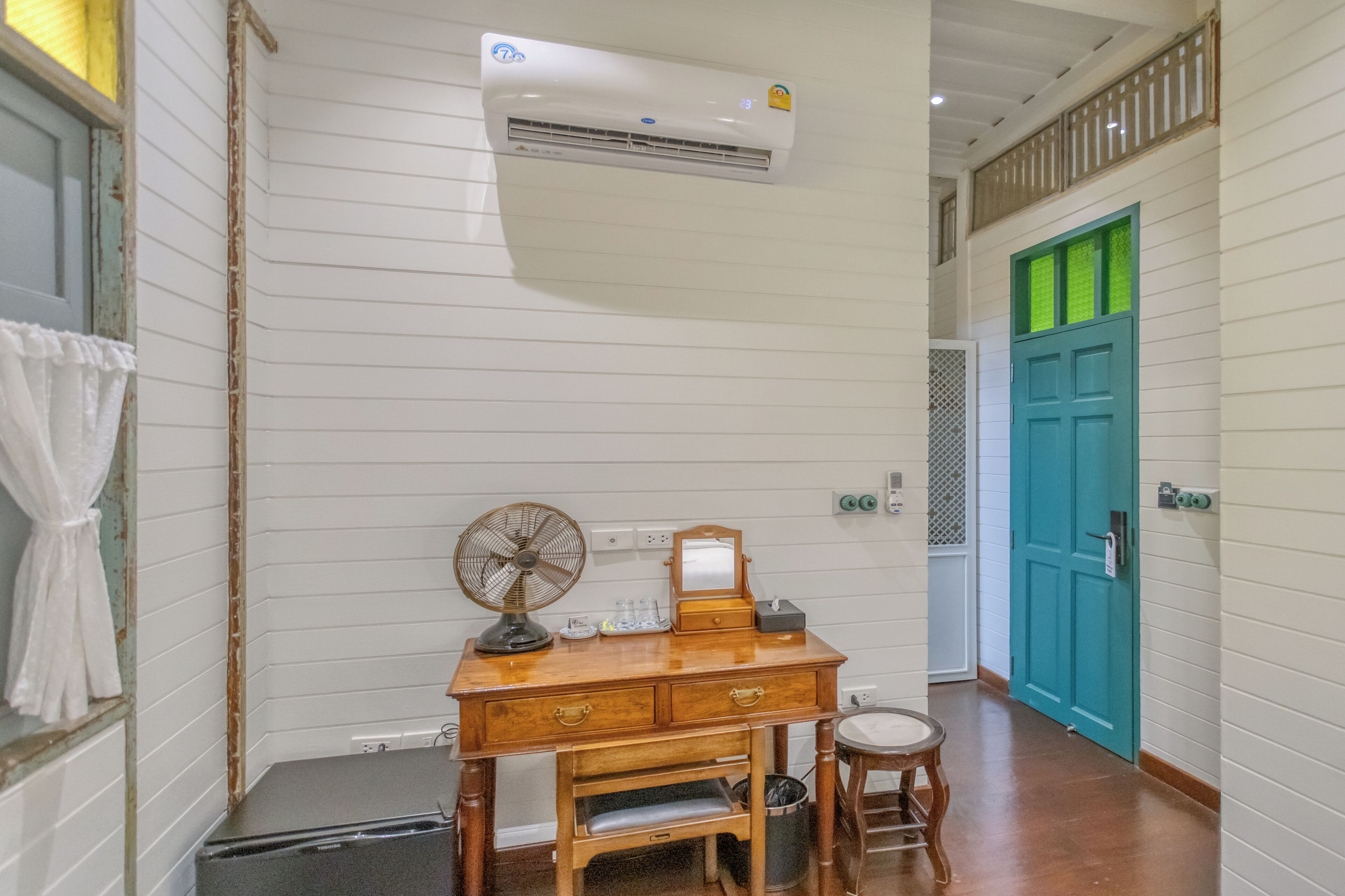Una habitación con un escritorio de madera, un ventilador, un espejo y una puerta verde azulada.
