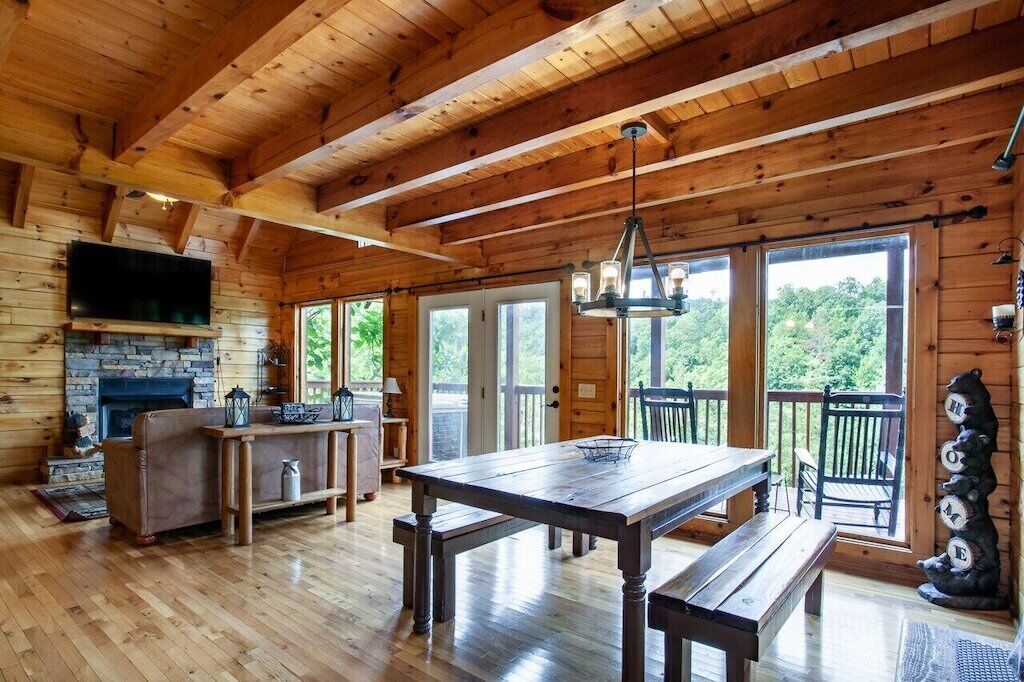 A wooden cabin interior with a stone fireplace, wooden dining table, benches, and large windows offering views of the outdoors.