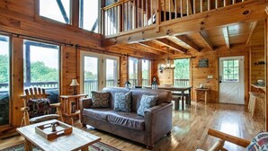 Een chaletinterieur met een houten vloer, een bank, een eettafel en een balkon met een leuning.