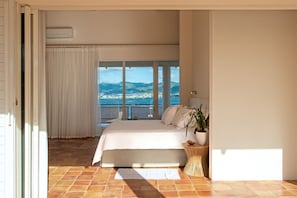 A bedroom with a large bed, a view of the sea and mountains, and a potted plant.