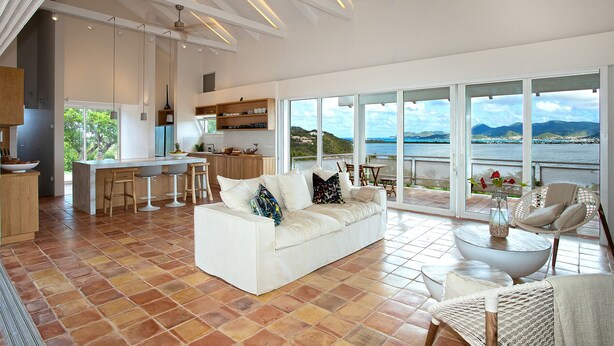 A spacious living area with a white sofa, a dining table, and chairs, a kitchen area, and large windows offering a view of the sea and hills. - Jasper -20% Low Season Promo | Ocean Front - Located in  Stunning Simpson Bay with Private Pool (Marigot)