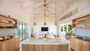 A spacious kitchen with wooden cabinetry, a central island, pendant lighting, and a large window providing a view of the outdoors. - Jasper -20% Low Season Promo | Ocean Front - Located in  Stunning Simpson Bay with Private Pool (Marigot)