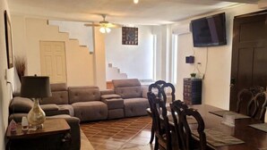 Una sala de estar con sofá, una mesa de comedor con sillas, un televisor montado en la pared y una escalera al fondo.