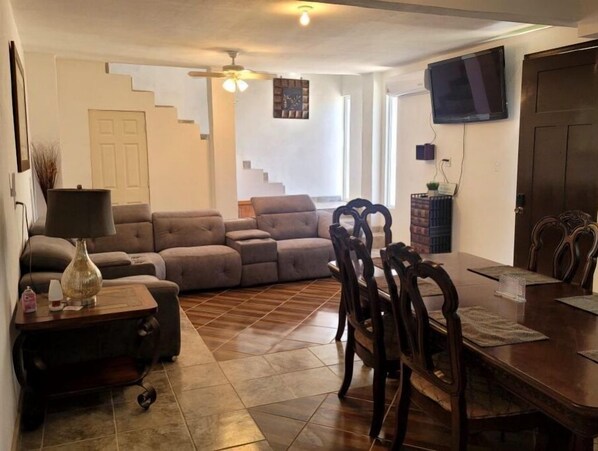 Uma sala de estar com sofá, mesa de jantar e cadeiras, televisão na parede e escada ao fundo.