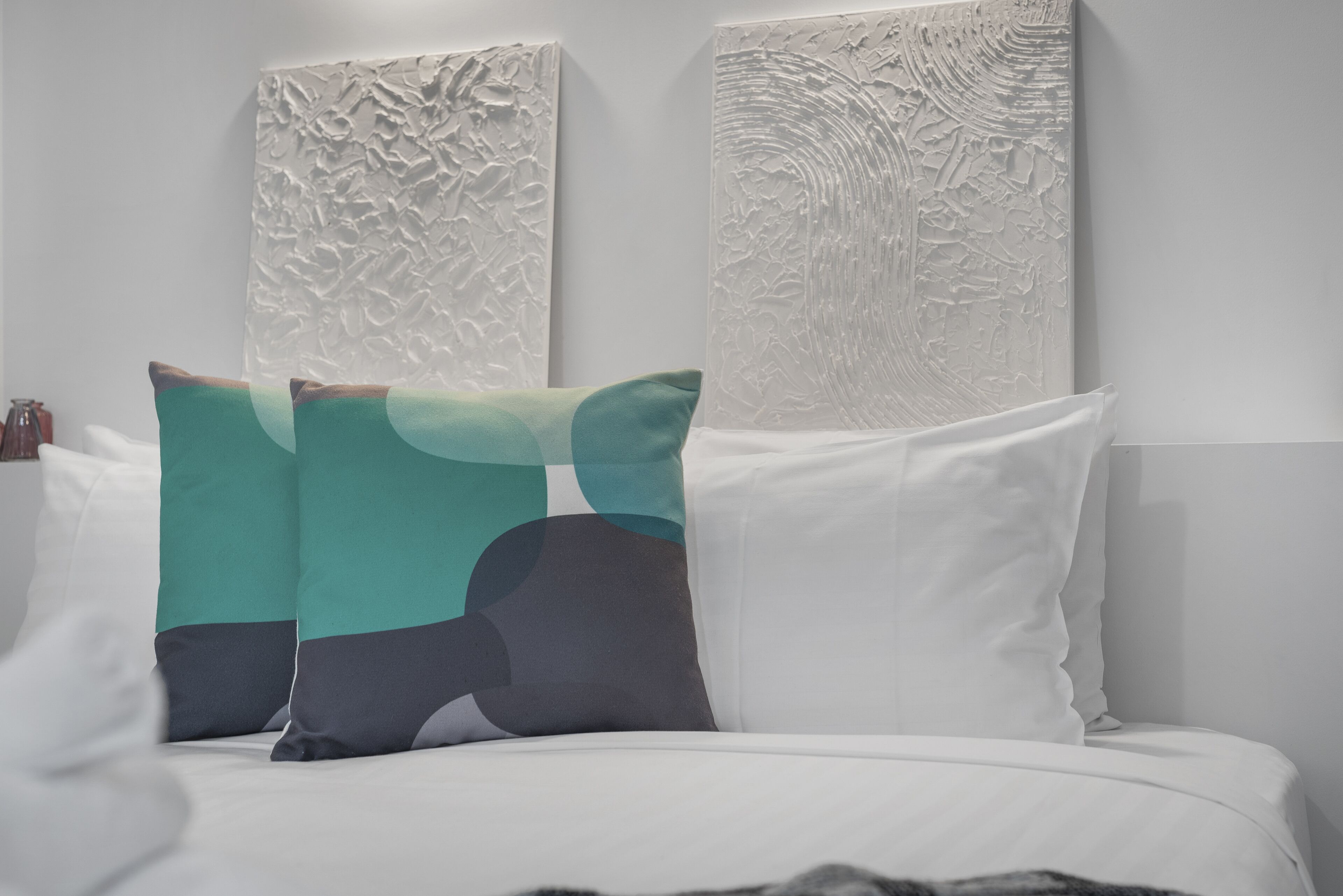 Una cama con ropa de cama blanca y almohadas verde azulado y grises, flanqueada por detalles decorativos en la pared.