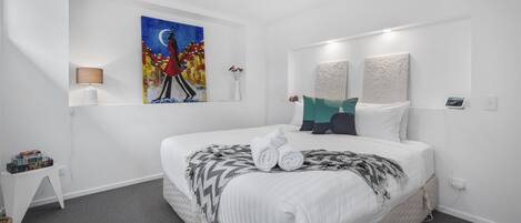 Una habitación de hotel moderna con una cama grande, un cuadro en la pared, una mesita auxiliar y una lámpara de noche.