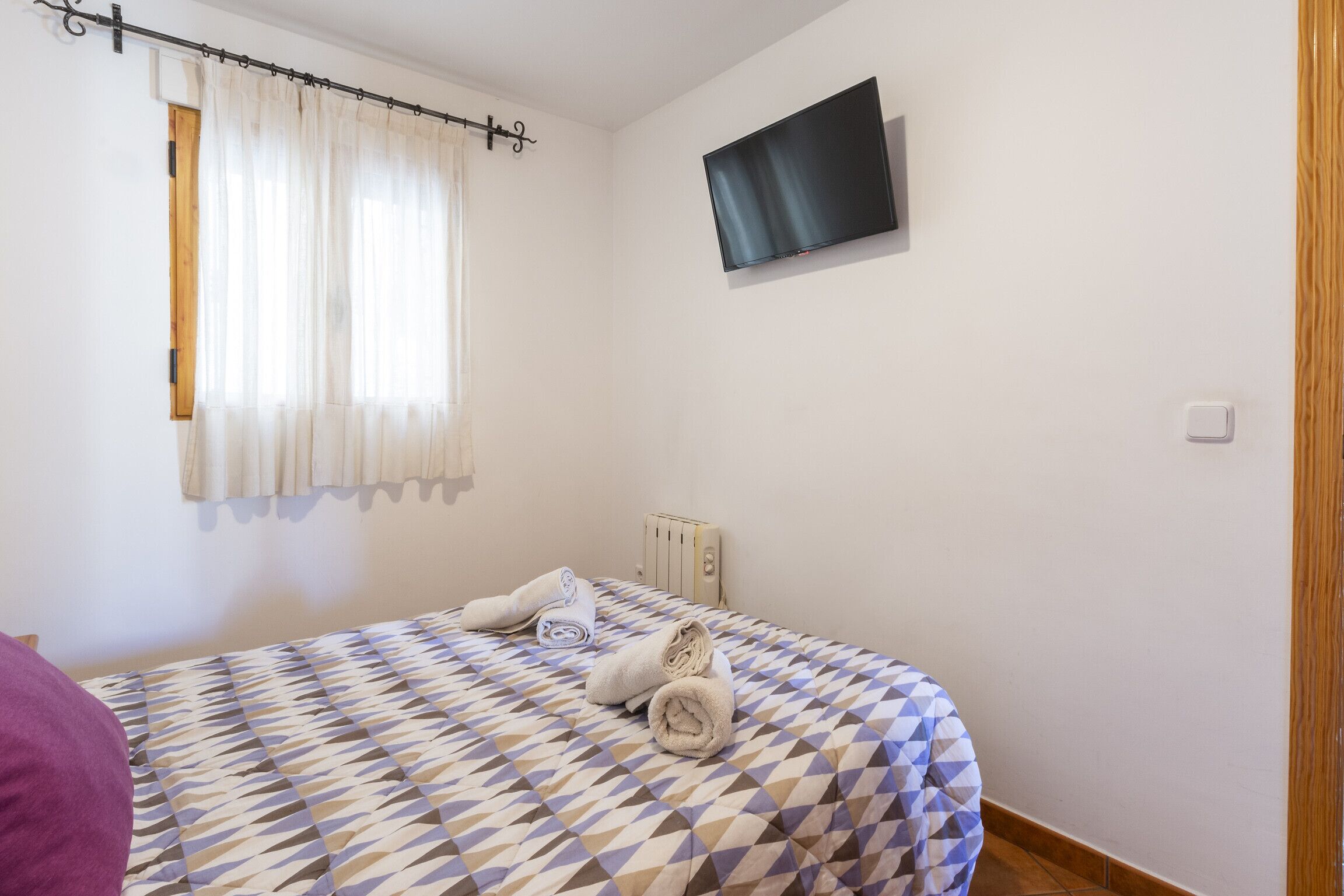 Quarto com colcha estampada, televisão de tela plana na parede, radiador e janela com cortinas.
