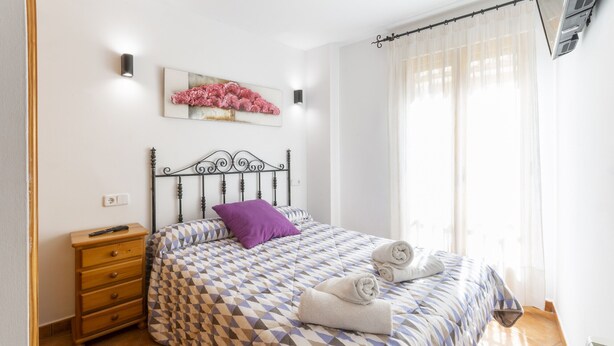 A bedroom with a patterned bedspread, a wooden nightstand, a purple pillow, and a wall-mounted artwork. - CARREBAIX II - Wonderful apartment with free WiFi. (Orba)