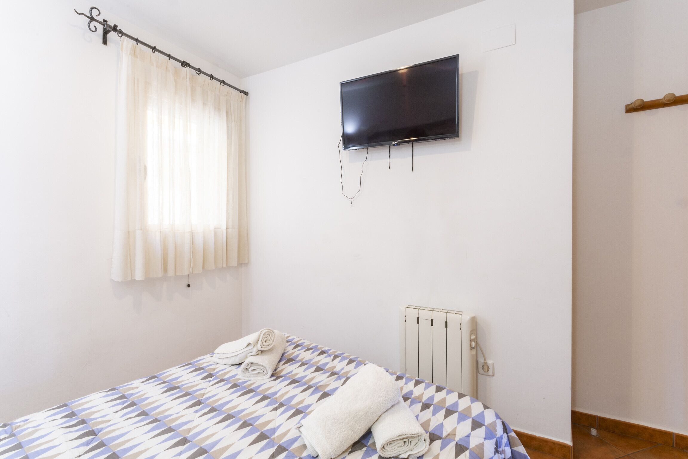 A bedroom with a bed, a TV mounted on the wall, a radiator, and a window with curtains.