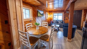 Interior acogedor de una cabaña con una mesa de comedor de madera, sillas blancas, un sofá y un televisor pequeño.