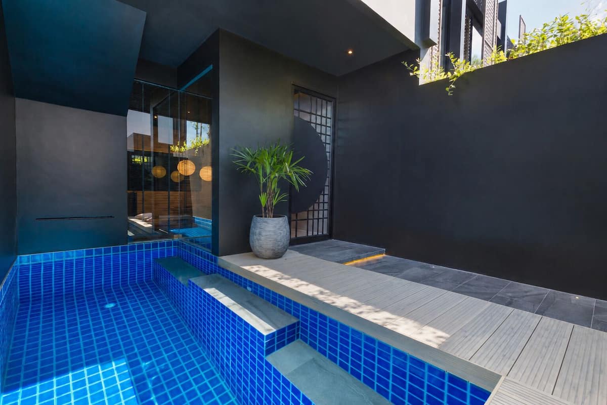 Un área de piscina moderna al aire libre con azulejos azules, una planta en maceta y una terraza de madera.