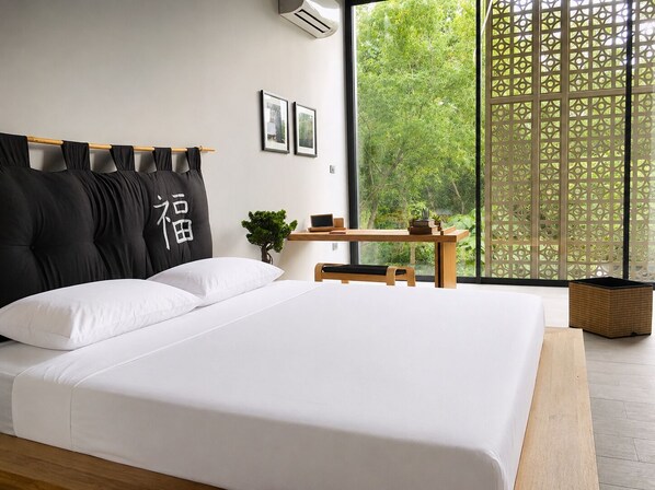 A modern bedroom with a large bed, a small desk, and a view of greenery through a window.