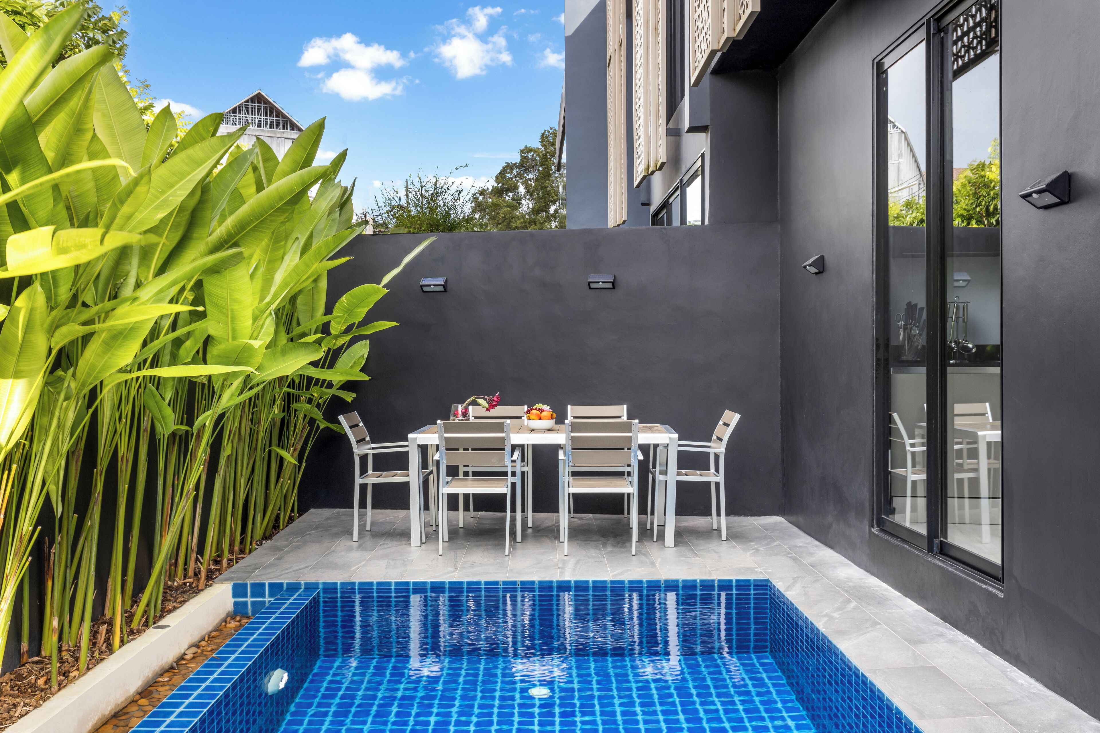 Un área de piscina moderna al aire libre con una mesa y sillas para comer, rodeada de plantas altas y un muro oscuro.