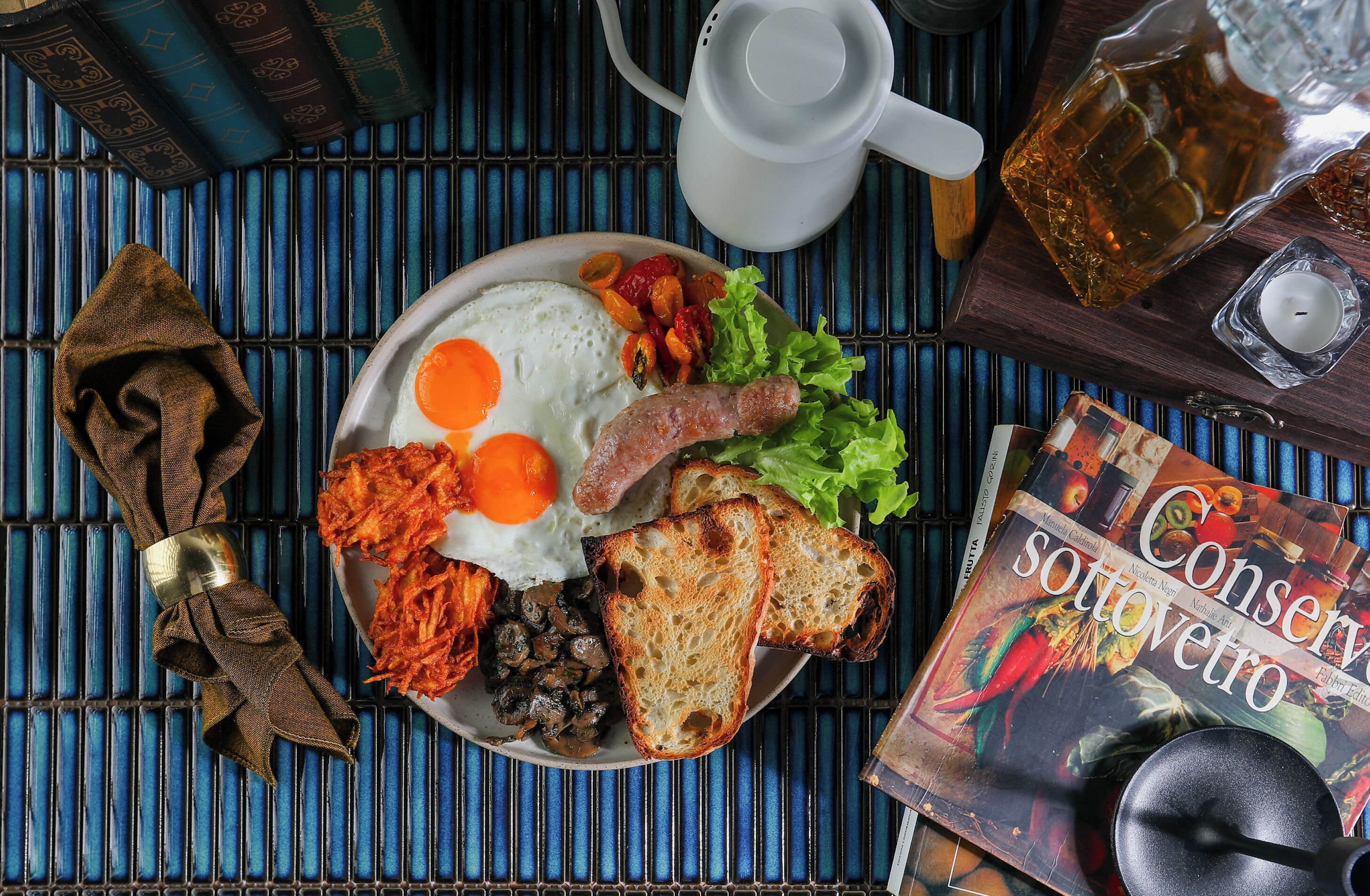 A plate of breakfast with eggs, sausage, mushrooms, and toast, a book titled 'Conservazione sotovetro', a teapot, and a candle on a blue mat.