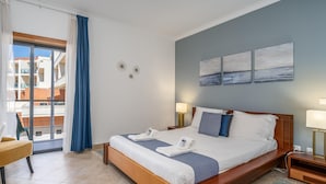 A hotel room with a large bed, a chair, a view of the outside, and artwork on the wall.