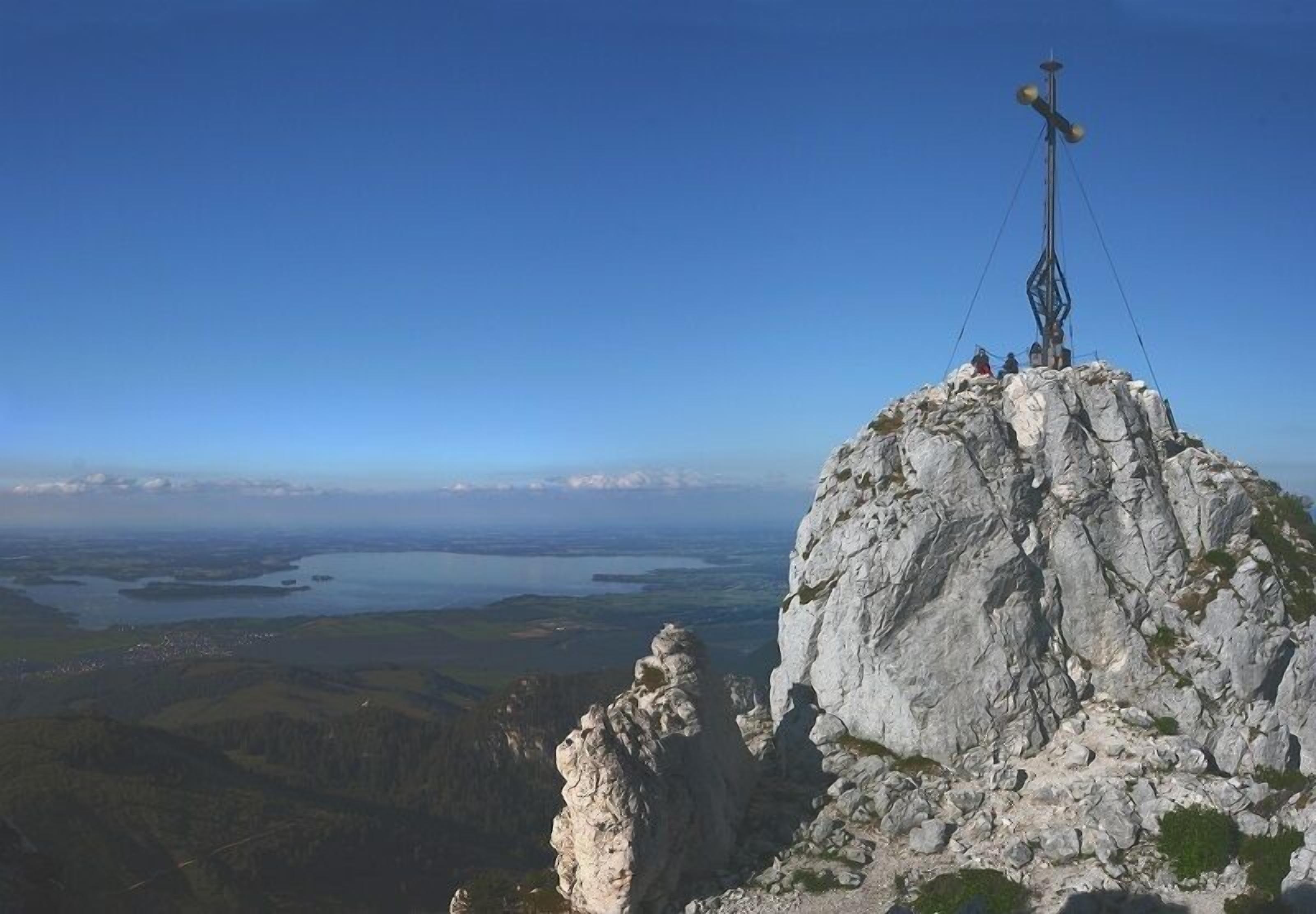 A rocky mountain peak with a cross structure on top, overlooking a valley with water bodies and distant towns.