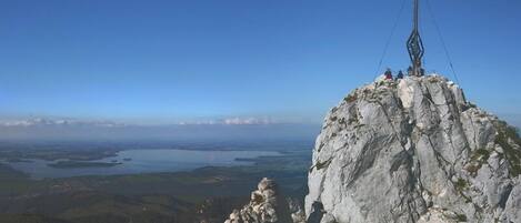 A rocky mountain peak with a cross structure on top, overlooking a valley with water bodies and distant towns.
